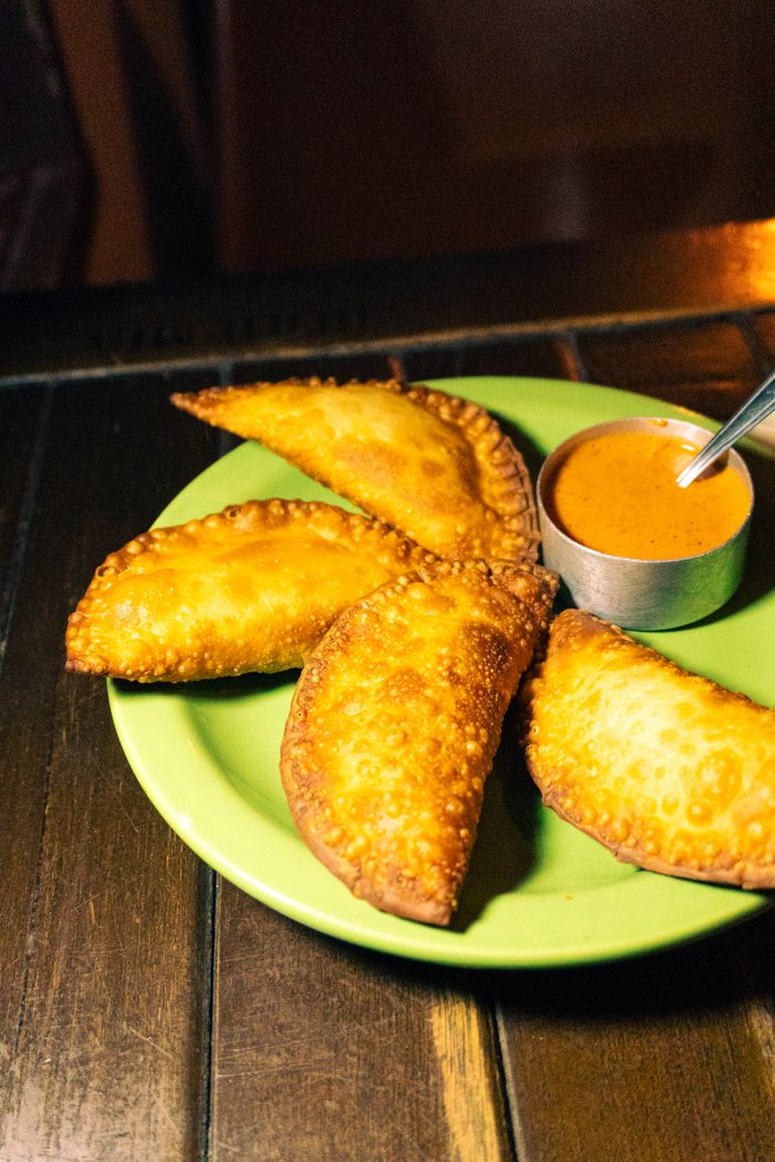 about-us-02 Four golden brown empanadas served with a tangy dipping sauce on a green plate.